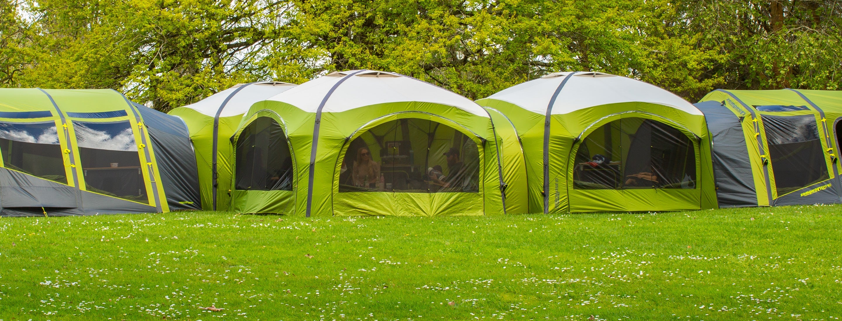Row of green tents with black accents on a grassy field with trees in the background