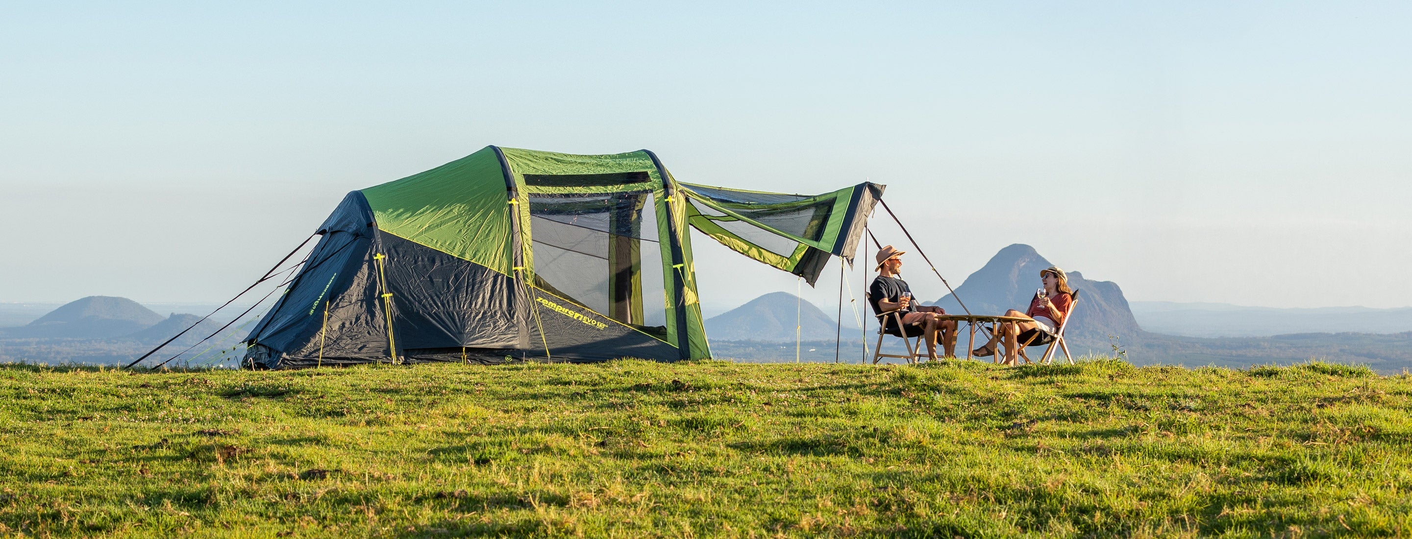 Two people sitting on chairs in front of a green tent in a grassy field with mountains in the background.
